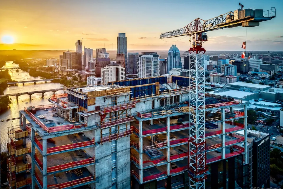 Large tower under construction with crane with American and Texas flags in downtown Austin, Texas against sunset sky.
